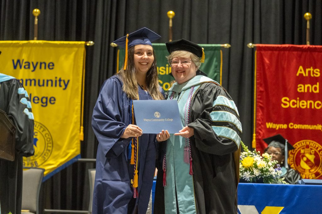 A graduate in a cap and gown receives her diploma from an individual also in a cap and gown.