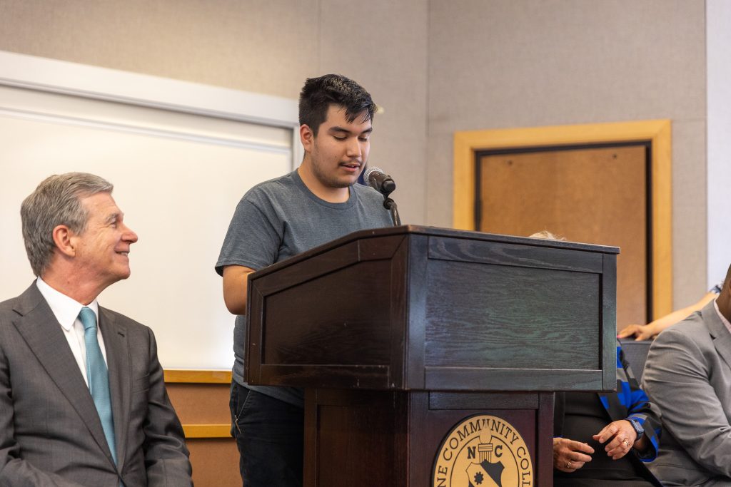 A young man stands in front of a podium and speaks in a microphone while another man sits and smiles.