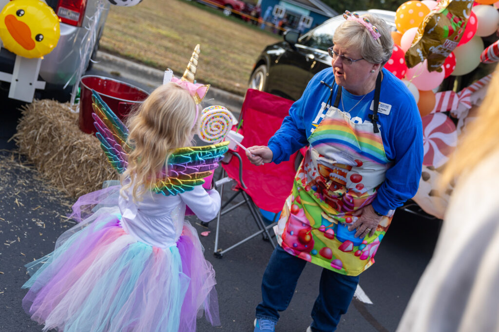 A woman in an apron holds a lollipop out to a child in costume.