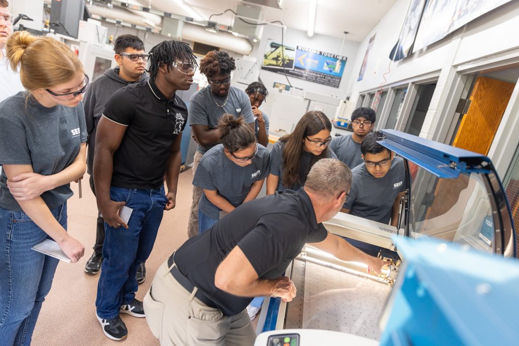 Students wearing safety glasses stand around a piece of equipment while a teacher points something out to them.