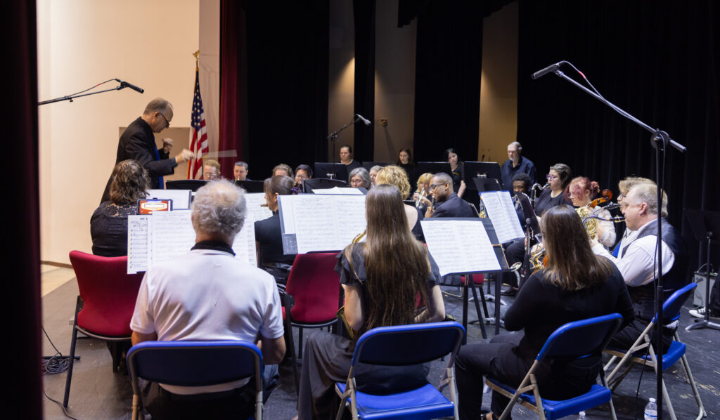 A group of people sit in a semicircle playing instruments while a man conducts them.