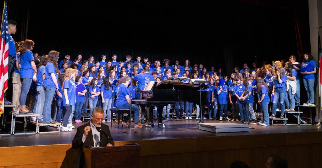 Students wearing blue shirts stand on a stage around a piano.