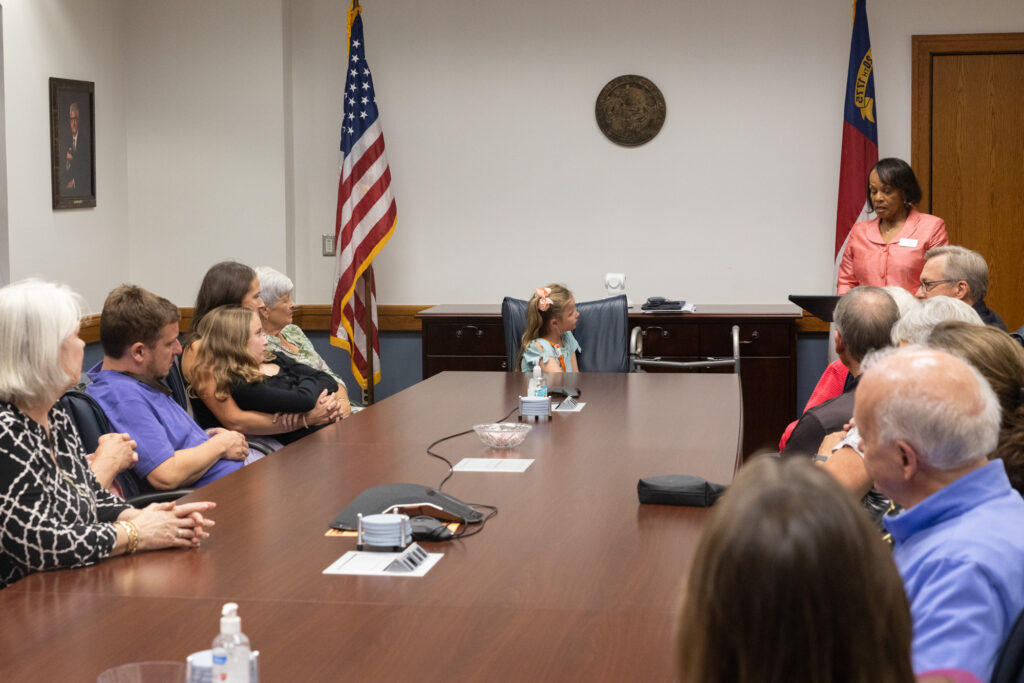 Individuals sit around a table while one stands and speaks at a podium.